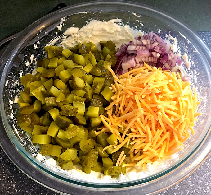 bowl of easy appetizer dip ingredients: dill pickles, cheddar cheese, red onion, cream cheese , and pickle juice