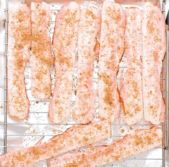 Brown sugar sprinkled over uncooked bacon strips on a wire rack on a baking sheet.