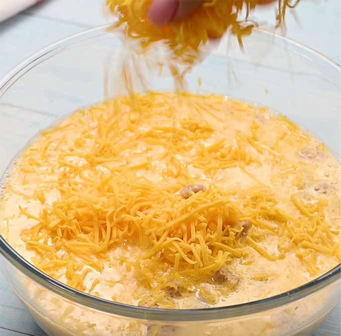 Shredded cheddar cheese being added to a mixing bowl of eggs, milk, salt, and pepper.