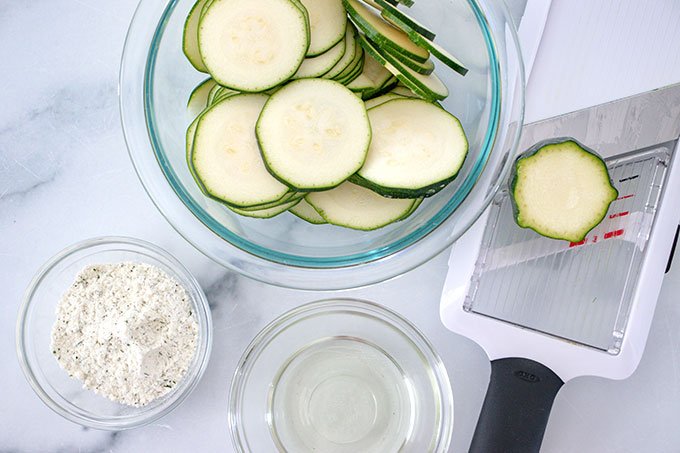 bowl full of thinly sliced zucchini pieces next to a mandolin and a bowl of ranch seasoning and vegetable oil