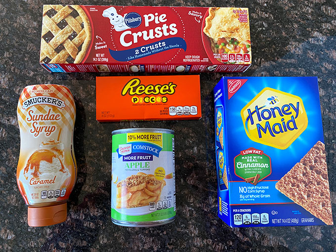 Ingredients for apple pie hack recipe:  pie crust, apple pie filling, Reeses pieces, caramel sauce, and cinnamon graham crackers