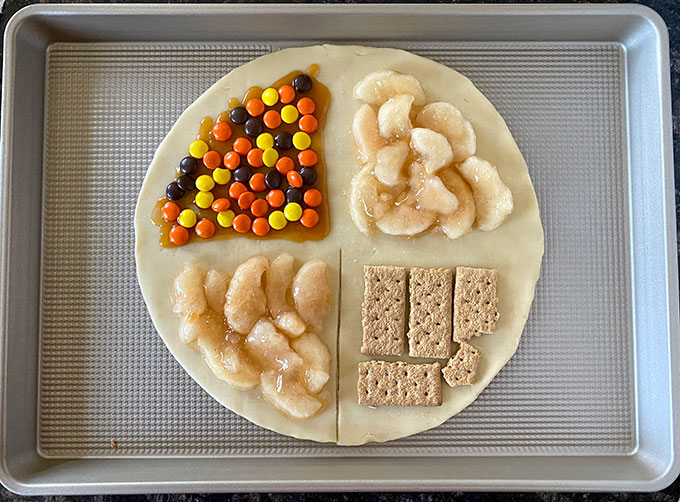 Pie crust with apple pie filling, caramel sauce, Reeses pieces, and cinnamon graham crackers in 4 sections.